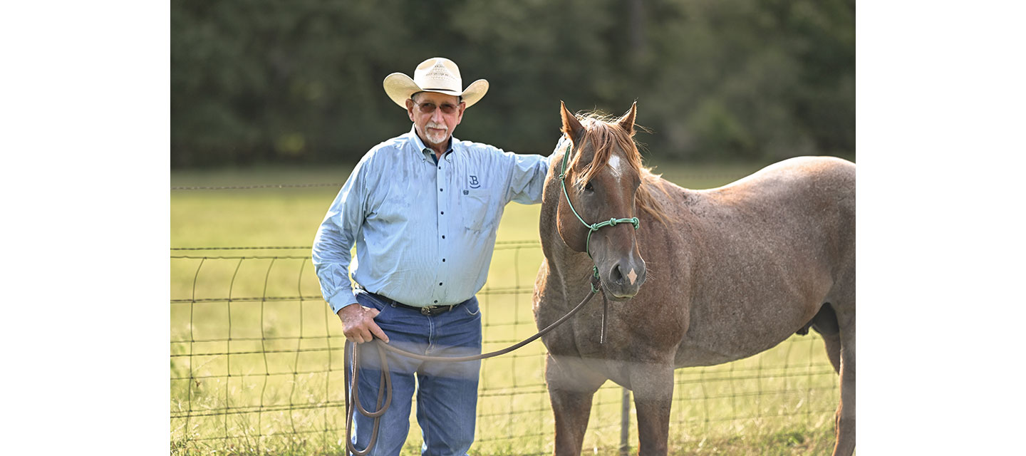 An older man in a cowboy hat and blue button-down shirt stands beside a roan horse in front of a wire fence.